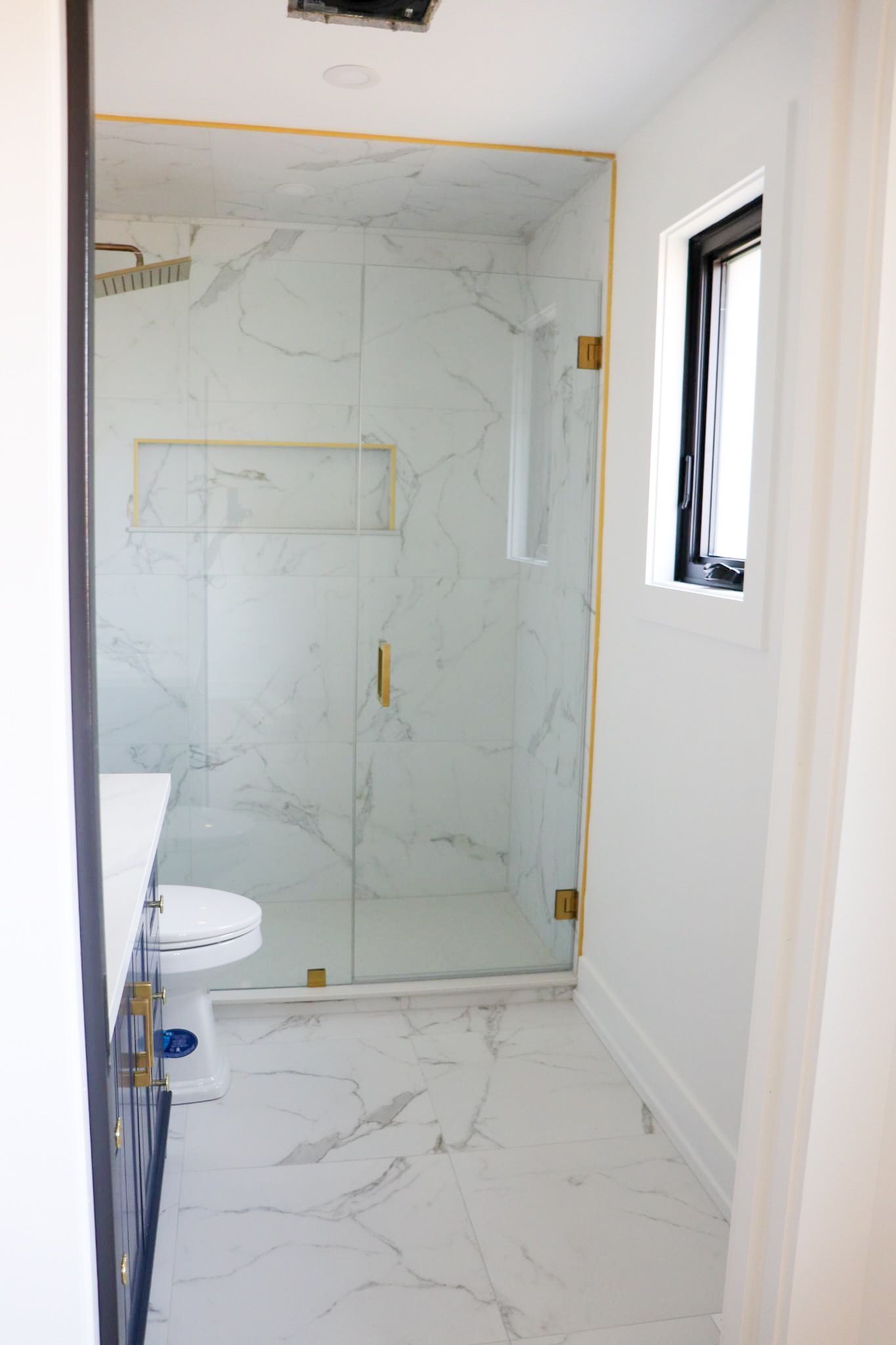 Bathroom with marble tile, gold hardware, glass shower, and a small black window.