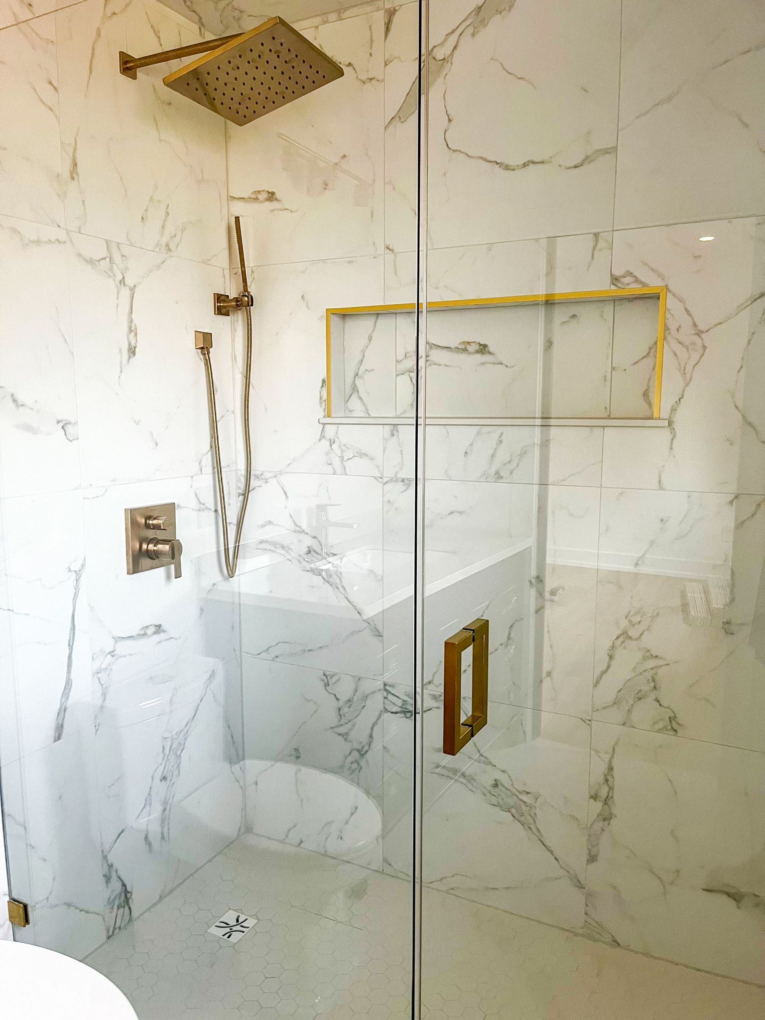 Gold-toned shower with white marble walls, glass door, and built-in shelf.