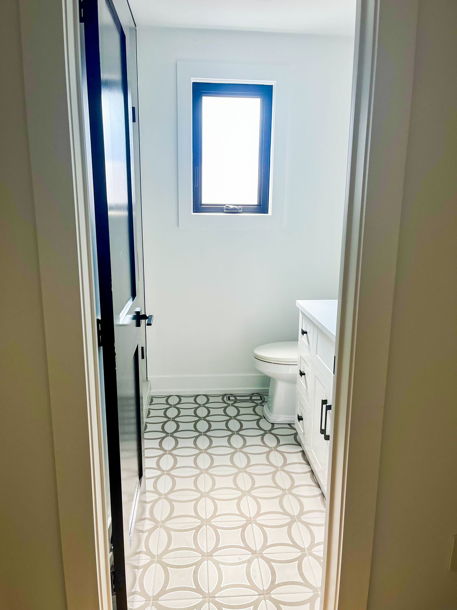 Bathroom with patterned floor, white walls, and a window. Toilet and vanity visible.