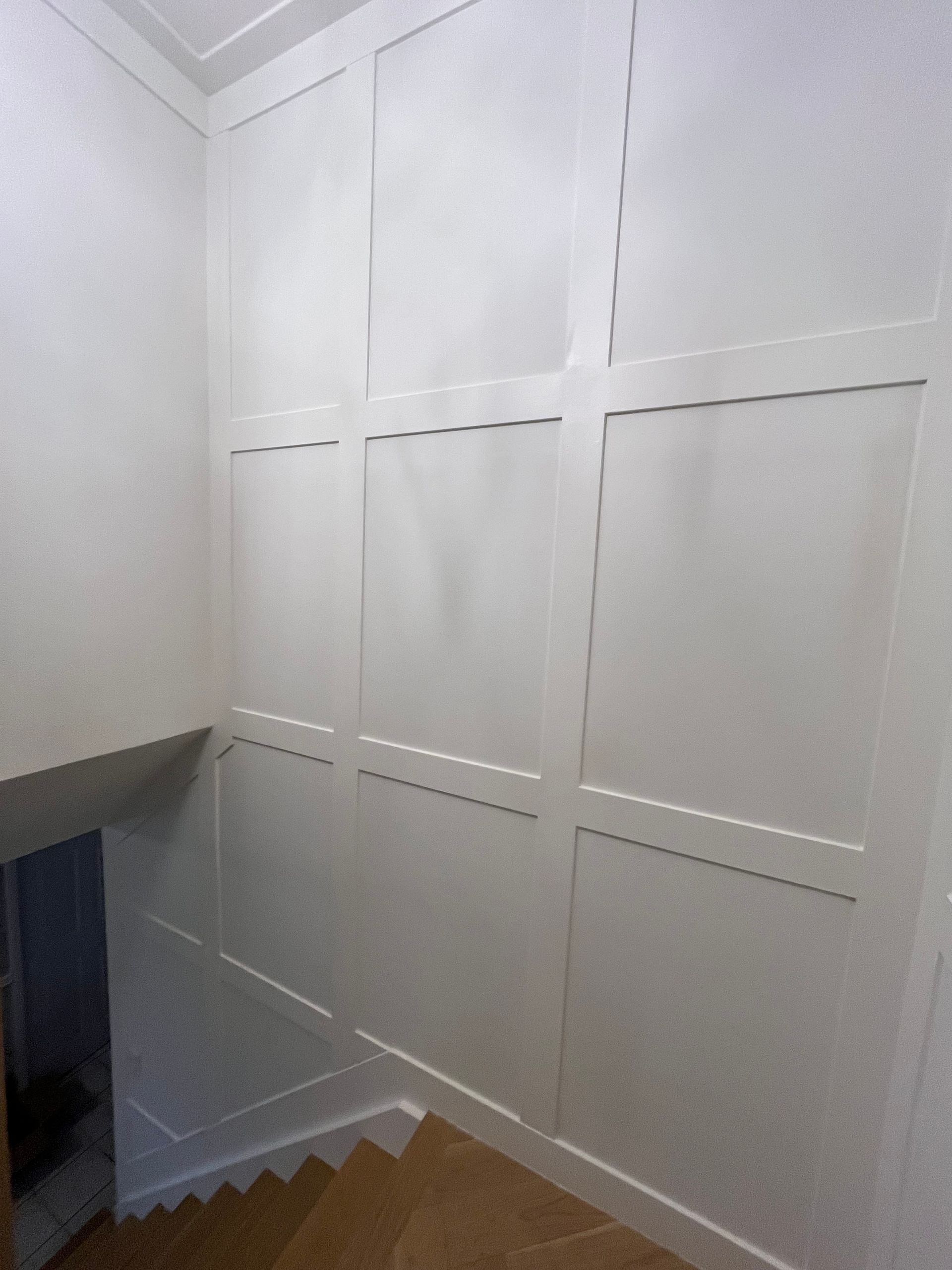 White paneled walls alongside a staircase. Wooden stairs and a dark opening are visible.