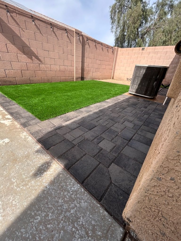 Backyard with artificial turf, dark paving stones, and a tan brick wall. A dark, angled object sits near the wall.