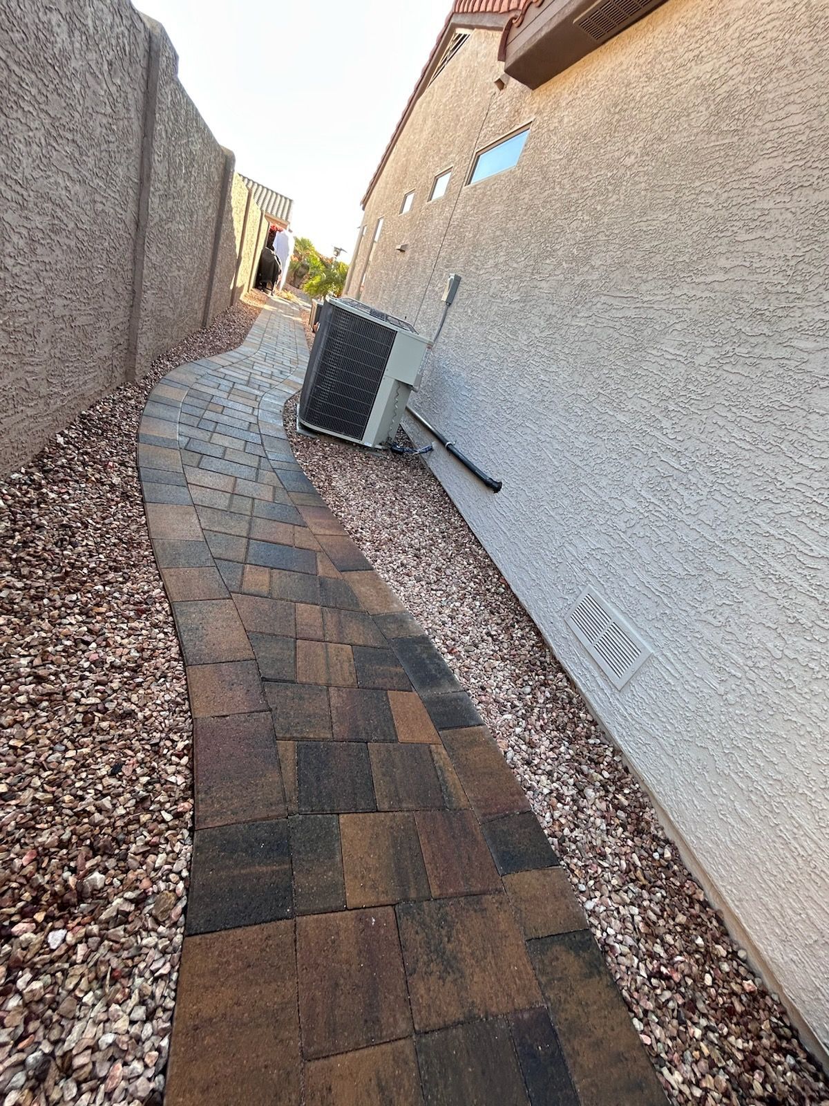 A narrow, winding pathway paved with brown bricks runs between tall beige walls and rocky landscaping.
