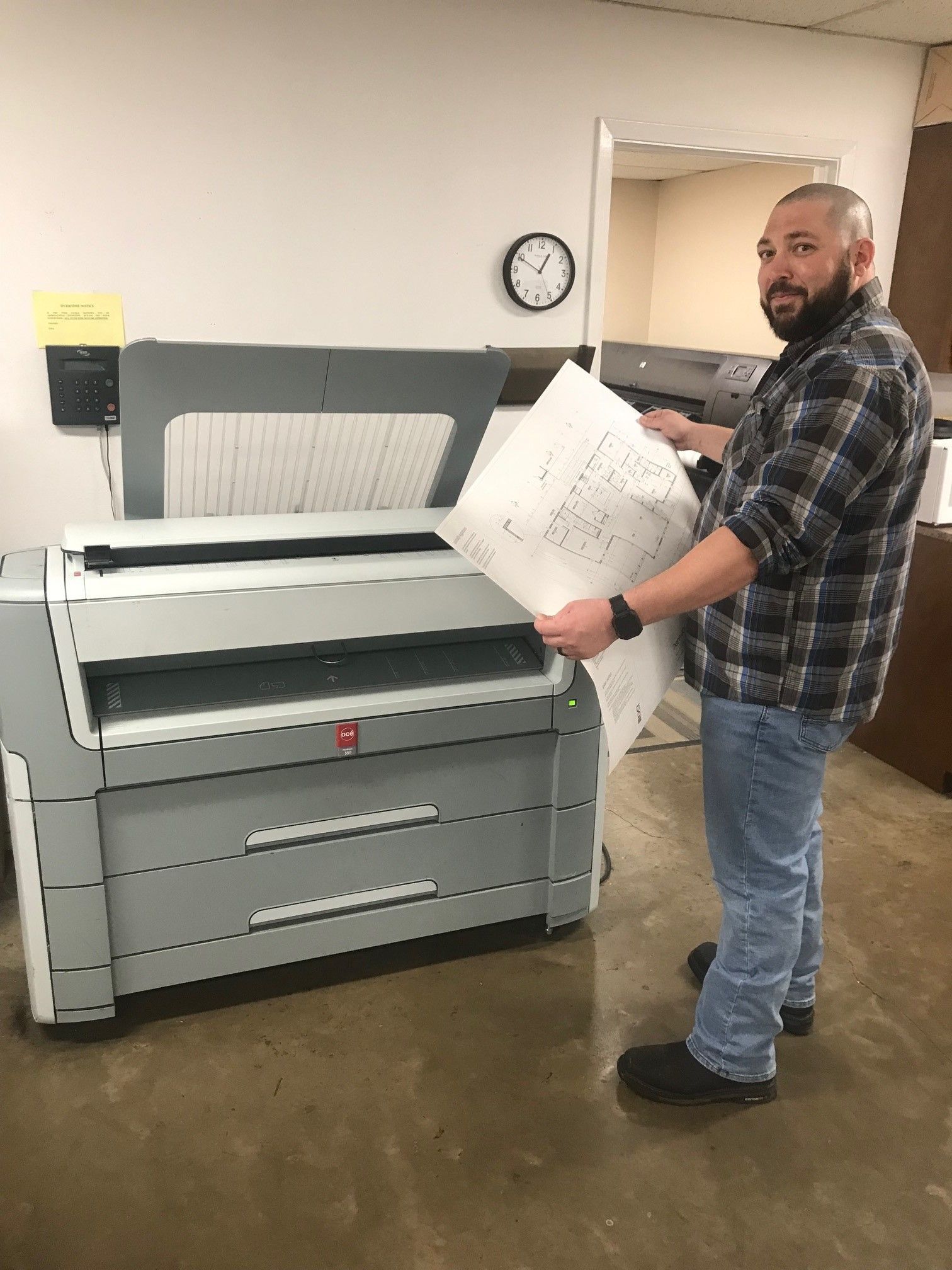 A man is standing in front of a large printer holding a piece of paper.