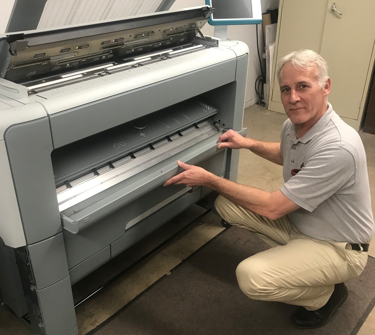 A man is kneeling down in front of a large printer.