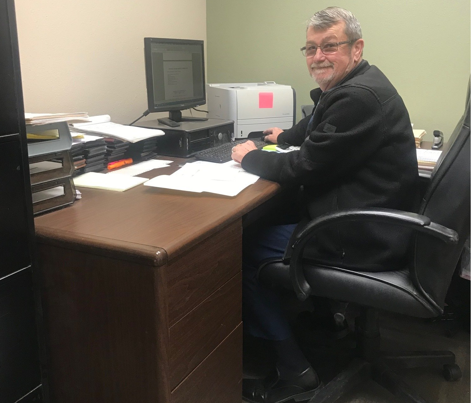 A man is sitting at a desk in front of a computer and printer.