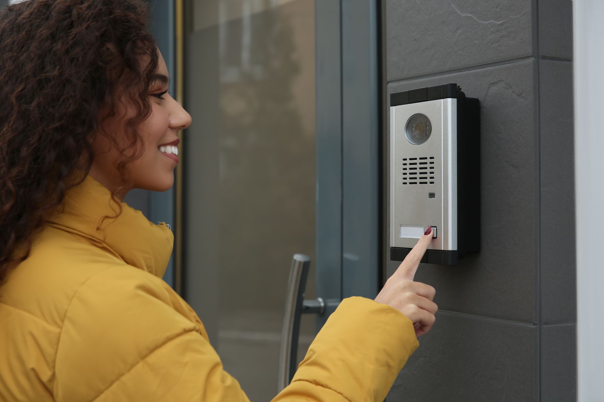 A person in a yellow jacket pressing a button on a modern silver door intercom system mounted on a gray wall.
