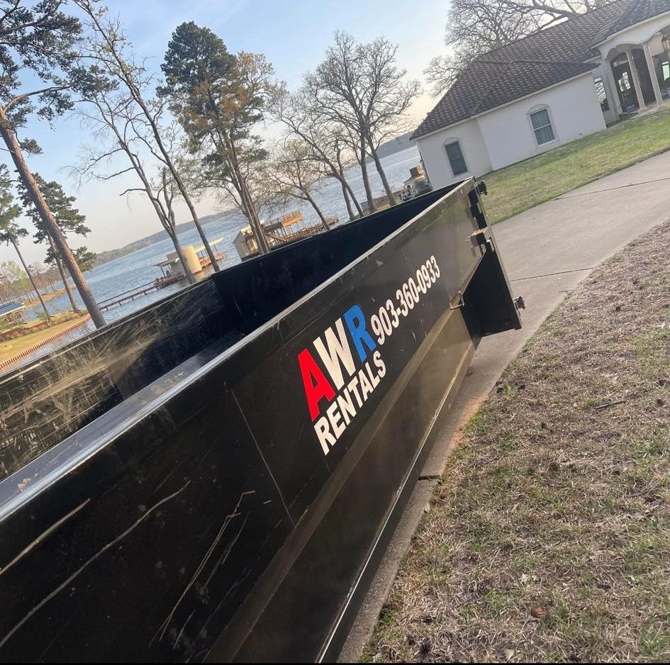 Black AWR Rentals dumpster on a grassy area near a lake and house.