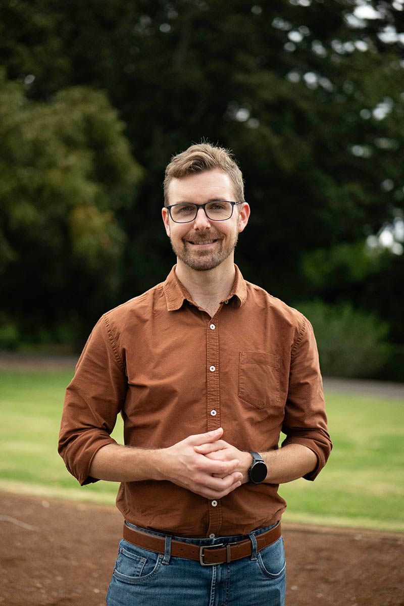 Man with glasses in brown shirt, hands clasped, stands in front of trees and a brown pathway.