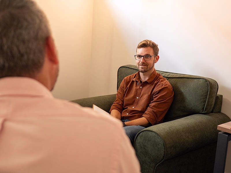 Man in orange shirt smiles, seated in a green armchair during a counseling session. Another person is seated opposite.