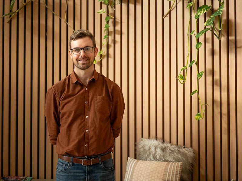 Man with glasses and brown shirt smiles in front of a wood-slat wall with plants.