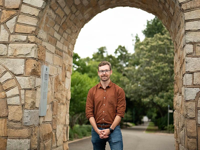 A person in a rust-colored button-down shirt and glasses stands under a stone archway outdoors.