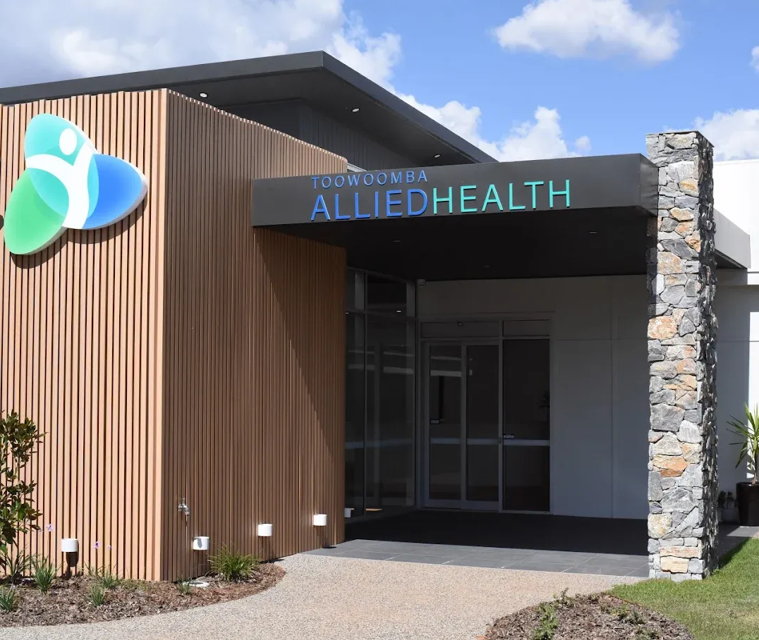 Allied Health building with blue, green logo. Wooden paneling, stone pillar, entrance under a dark awning.