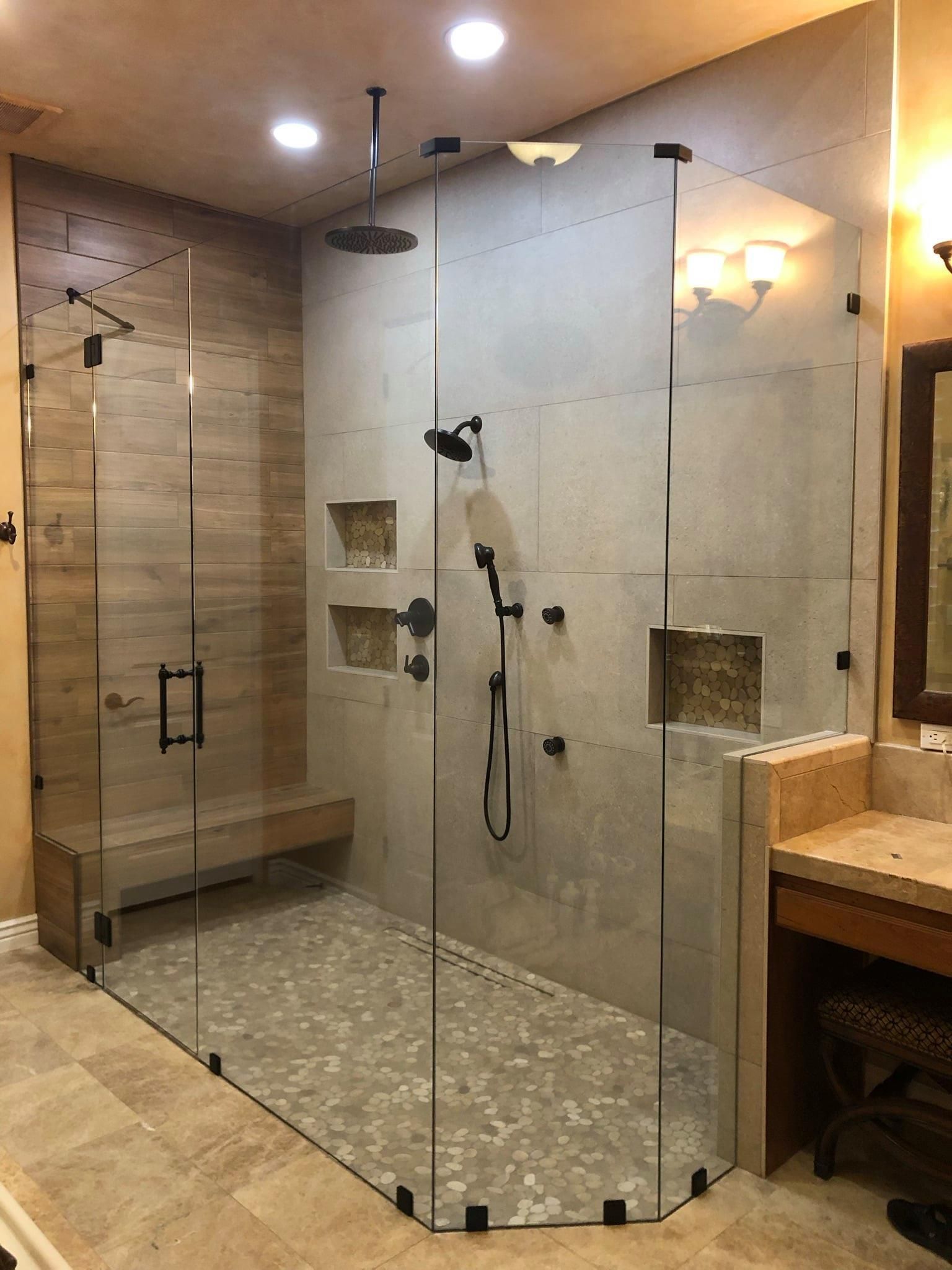 Modern shower with glass walls, pebble floor, built-in bench, and multiple showerheads. Beige tile and recessed shelves.