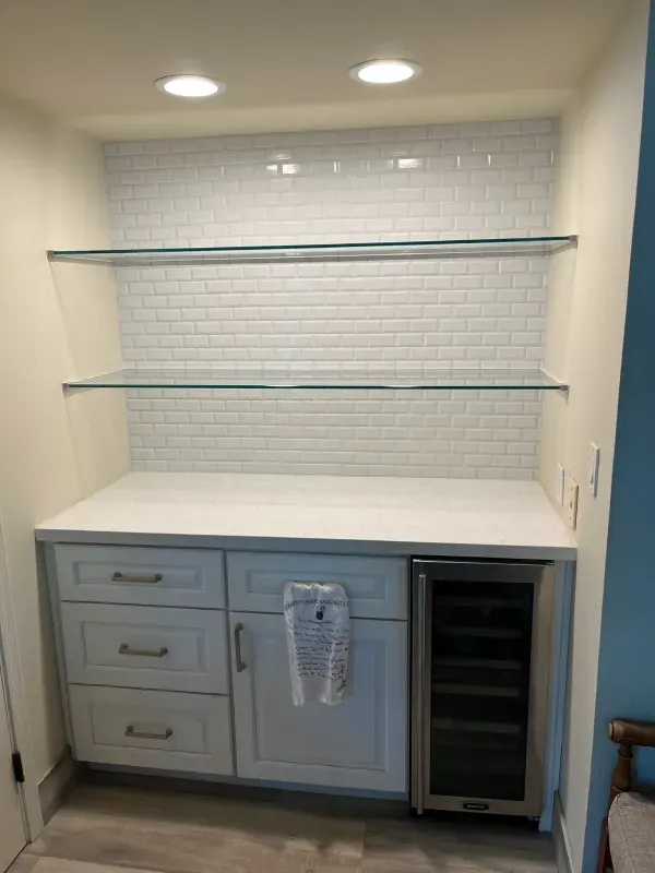 A kitchen with white cabinets , glass shelves and a wine cooler.