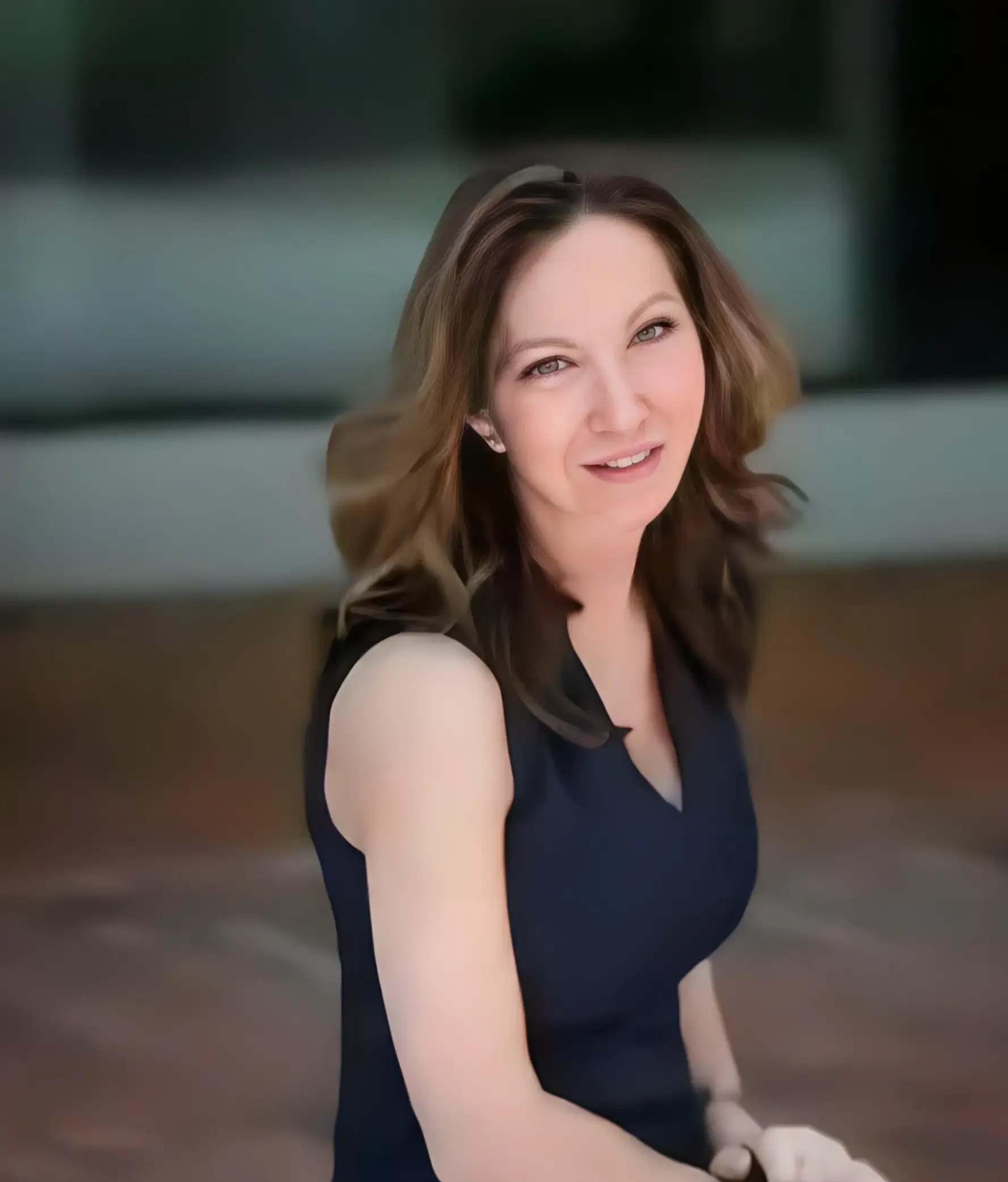 A person with shoulder-length wavy brown hair wears a sleeveless dark blue top, smiling while looking at the camera.