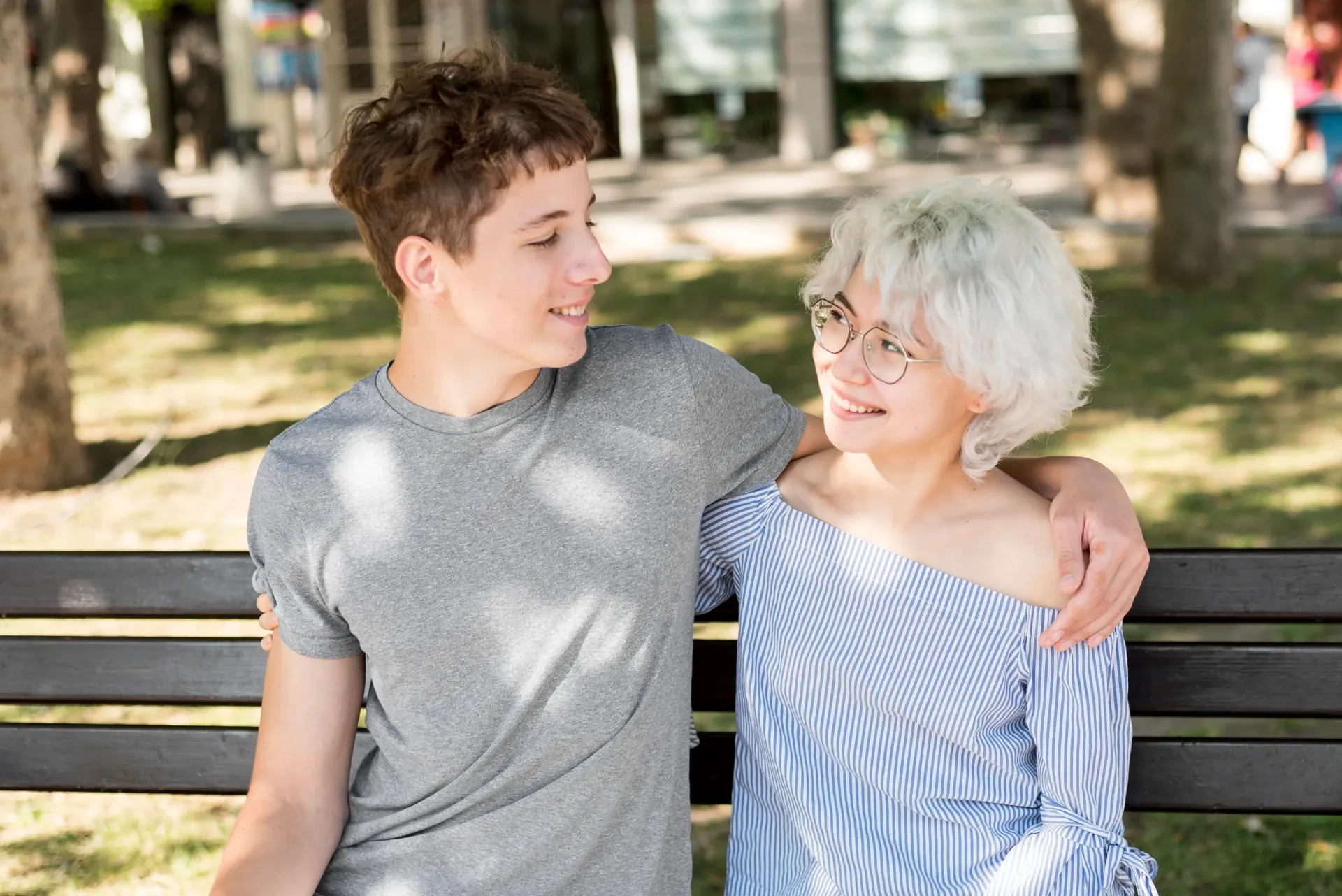 Two people with short hair sitting on a park bench, looking at each other and smiling, with arms around one another.