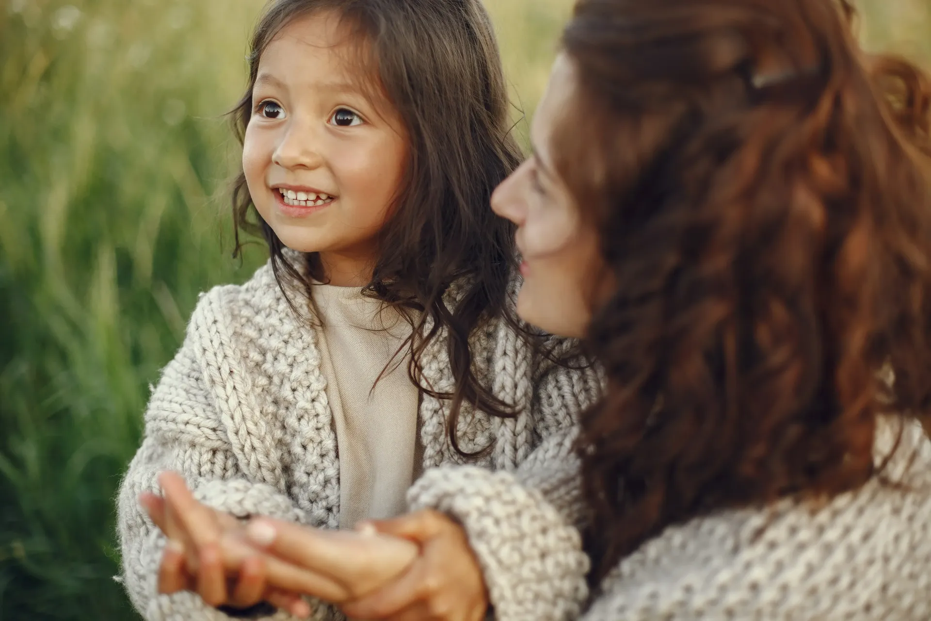 A person and a child in matching knit sweaters hold hands while looking off into a grassy field.