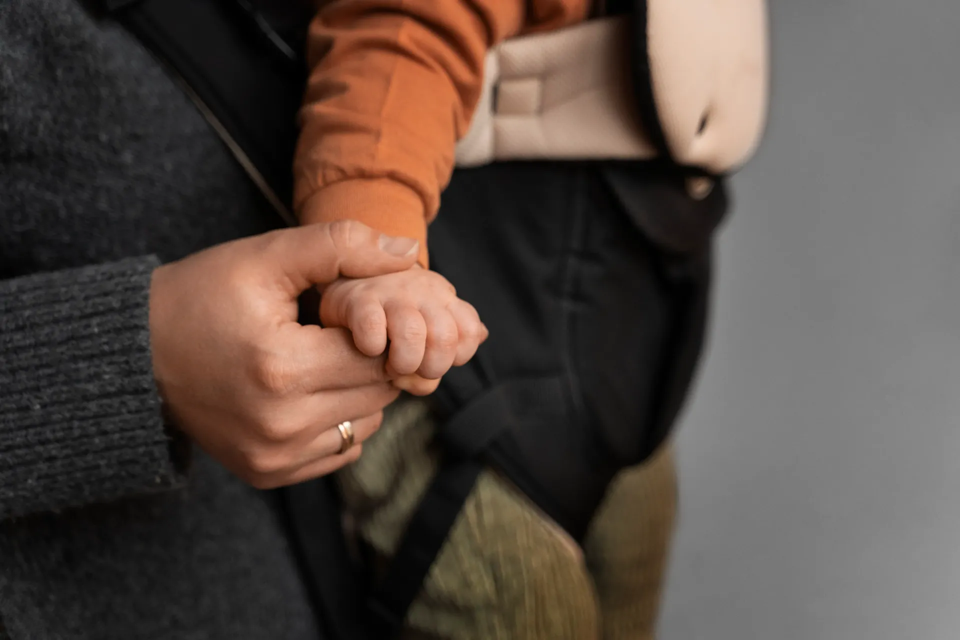 An adult hand in a grey sweater holds the small hand of a child wearing an orange sleeve in a baby carrier.