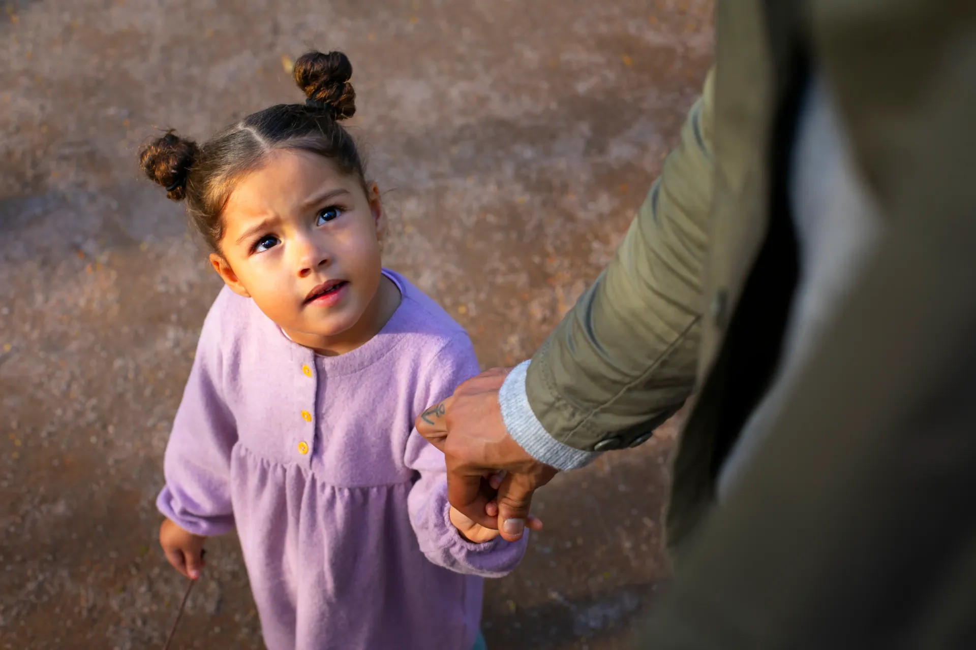 A child in a lavender dress looks up while holding the hand of an adult wearing an olive green jacket.