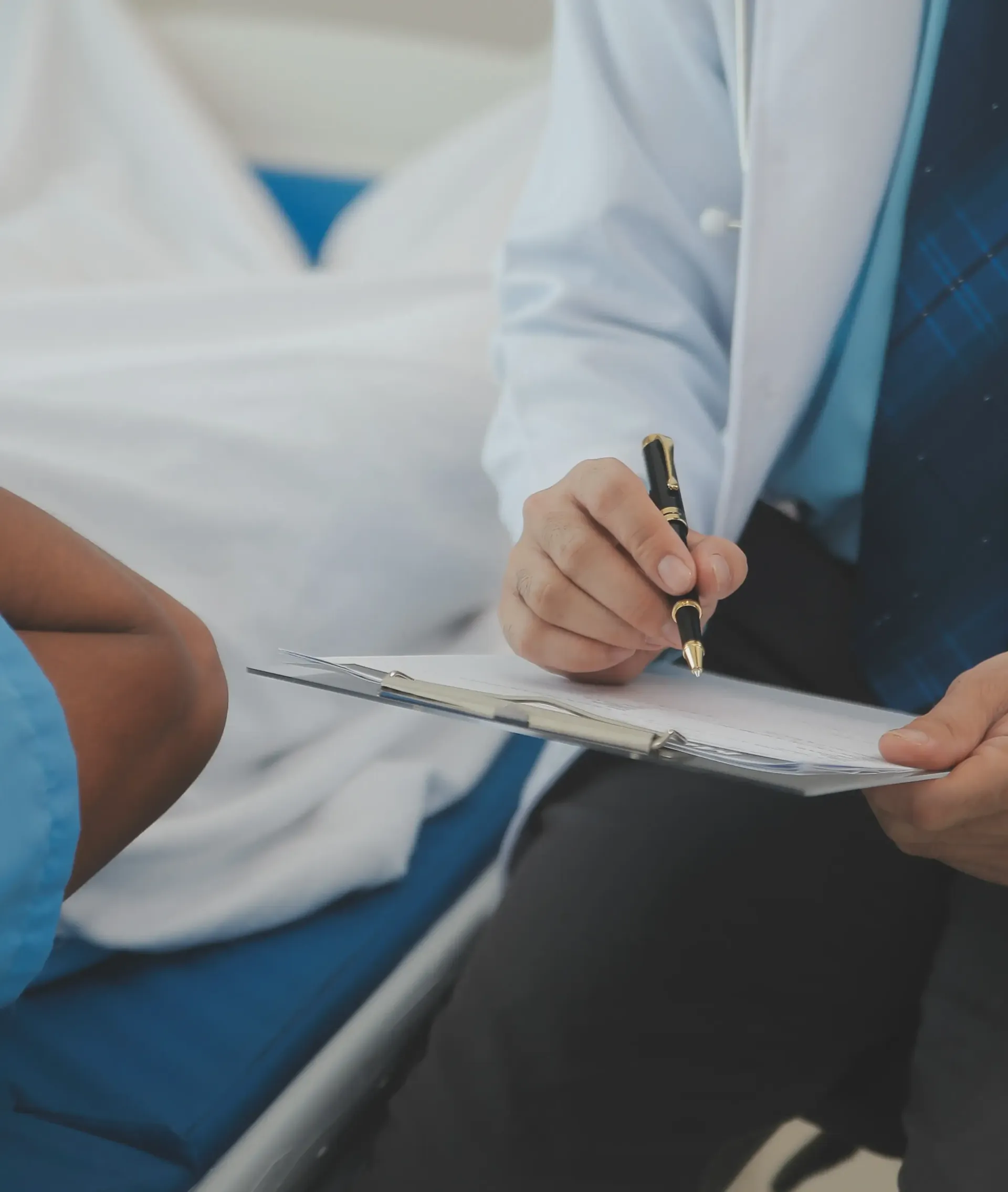 A medical professional in a white coat writes on a clipboard next to a patient lying in a hospital bed.