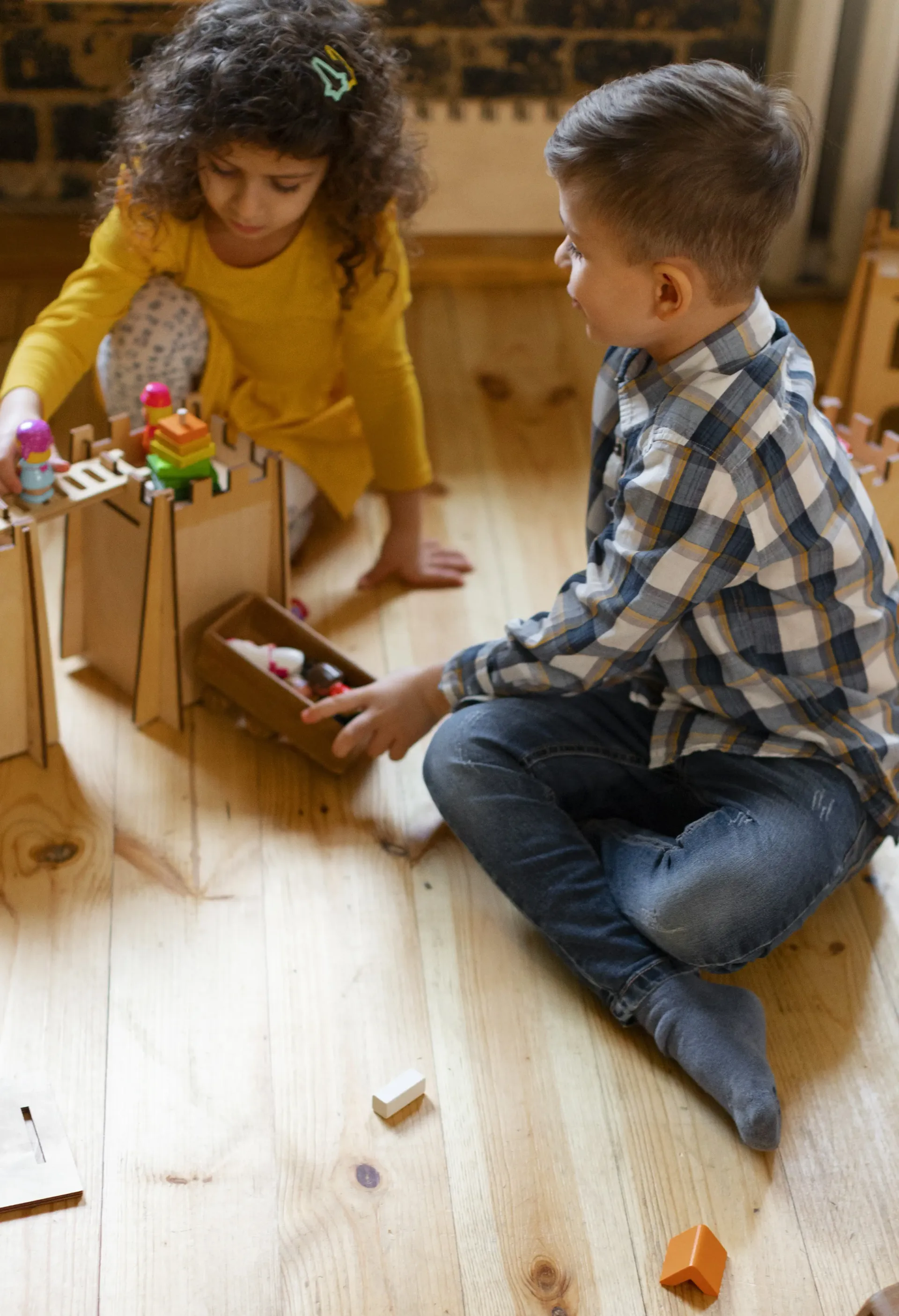 Two children sitting on a wooden floor, playing with a wooden castle and colorful block toys.