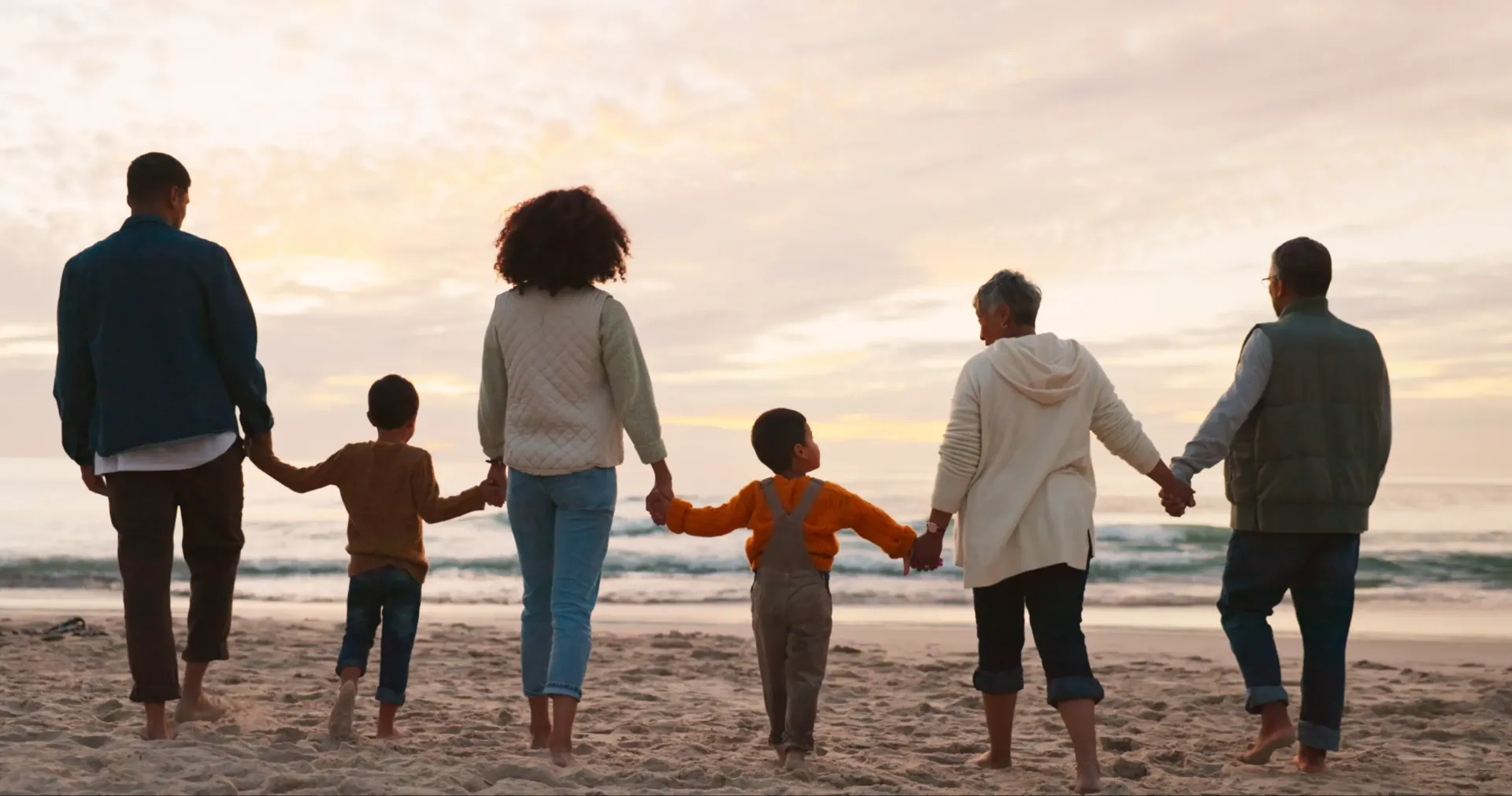 Six people stand in a line on a sandy beach holding hands and facing the ocean during sunset.
