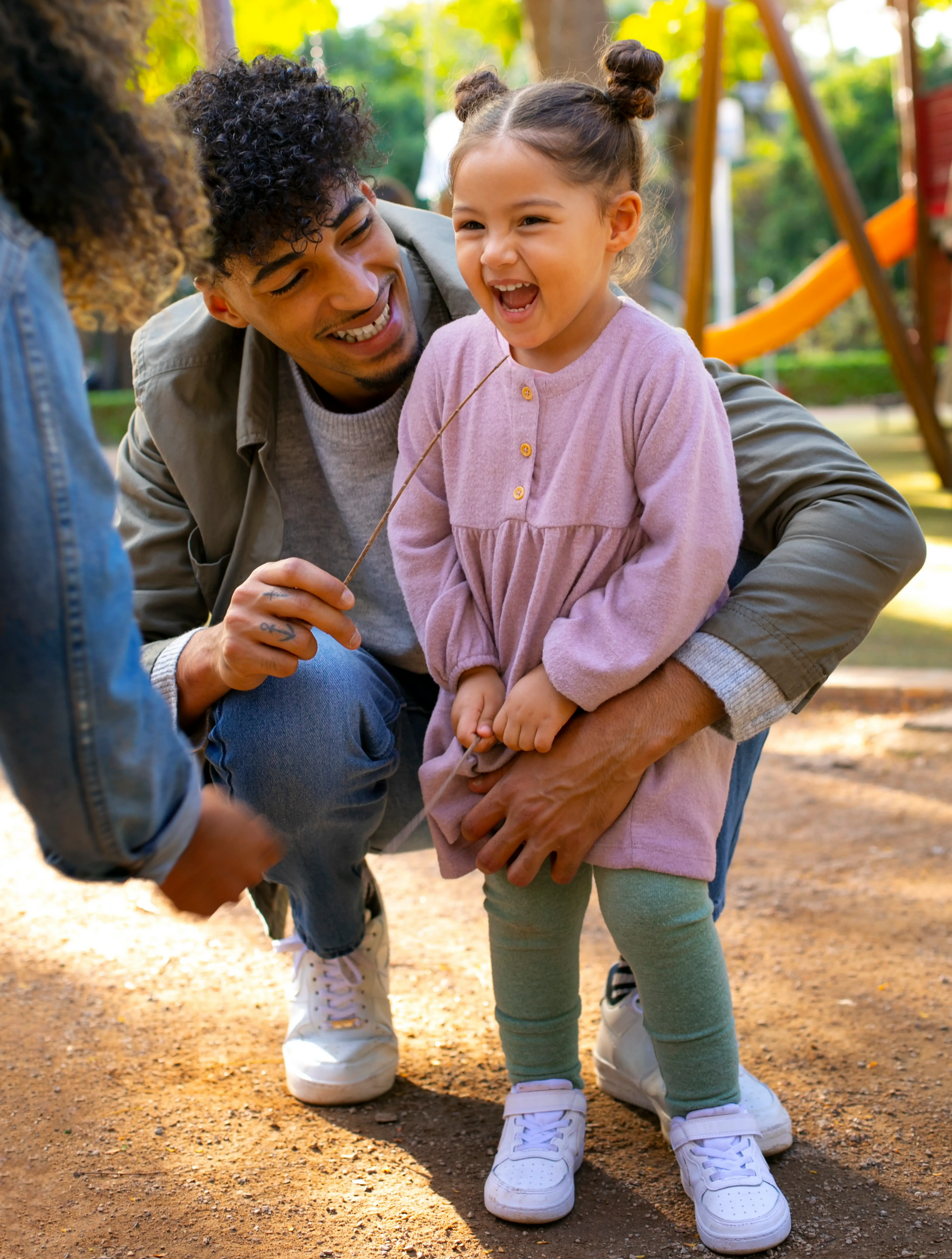 A smiling child in a pink dress and green leggings stands outside while a parent crouches behind them, holding a twig.