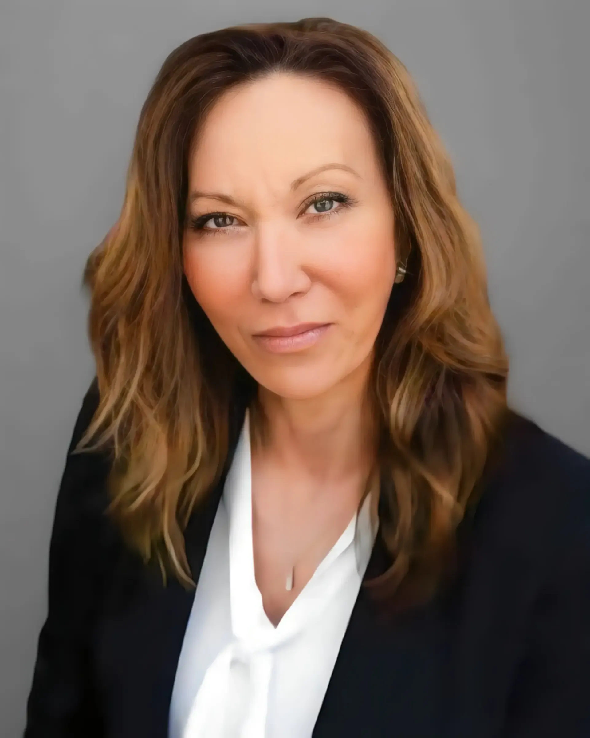 Headshot of a person with shoulder-length wavy brown hair, wearing a white blouse and dark blazer against a grey backdrop.