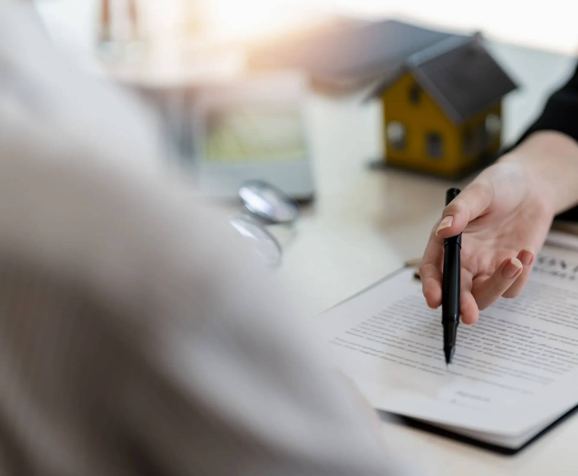 A person points a pen at a document on a desk next to a miniature house model, suggesting a real estate consultation.