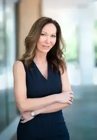 A professional portrait of a person with shoulder-length brown hair, wearing a dark sleeveless top, arms crossed, blurred.