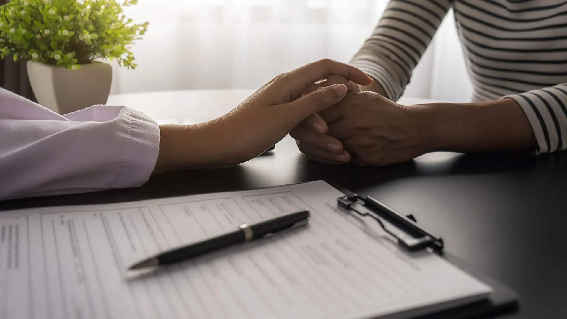A professional comforting a person by holding their hands over a document and pen on a table, with a potted plant nearby.