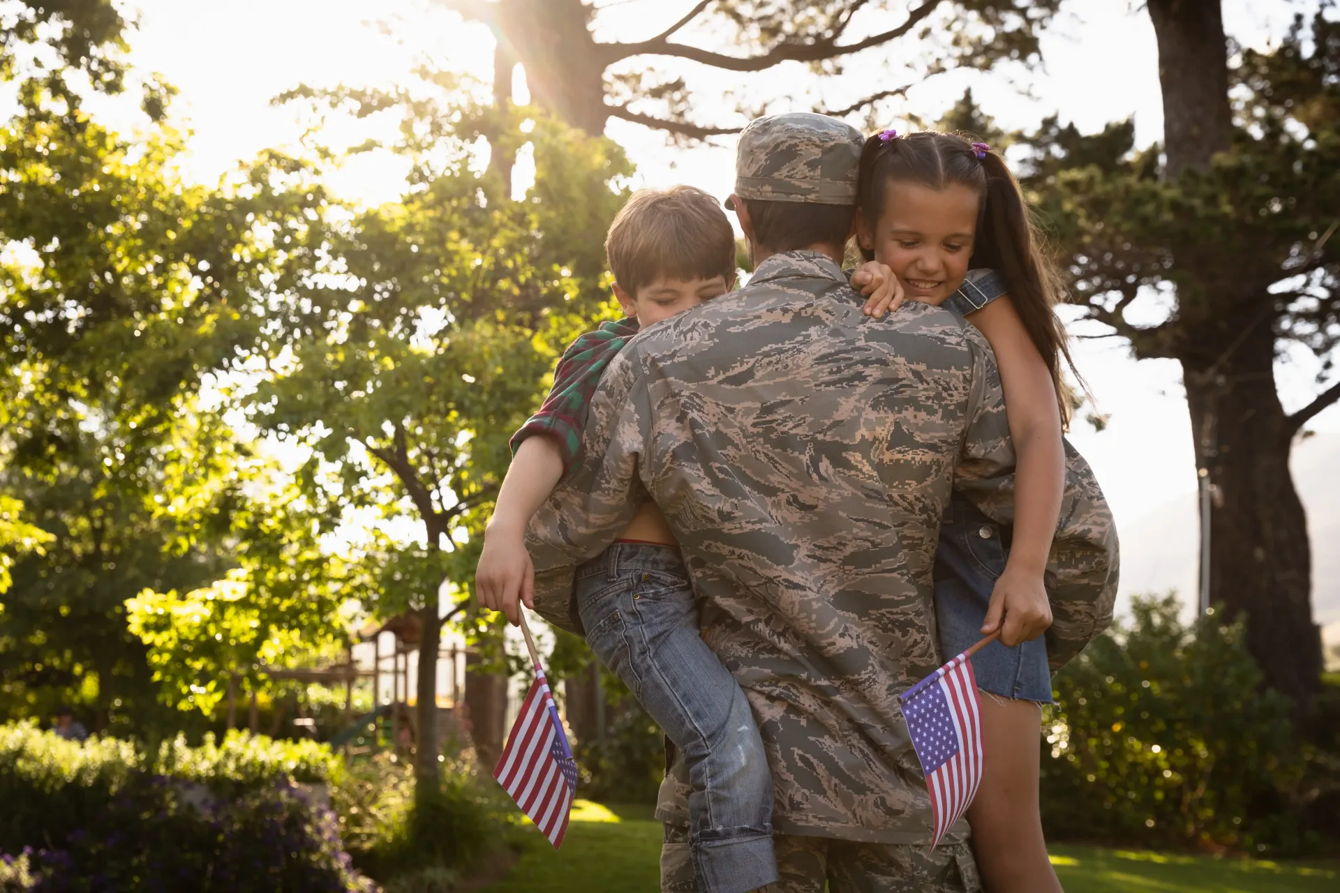 A military member in uniform embraces two children holding small American flags in a sunlit outdoor setting.