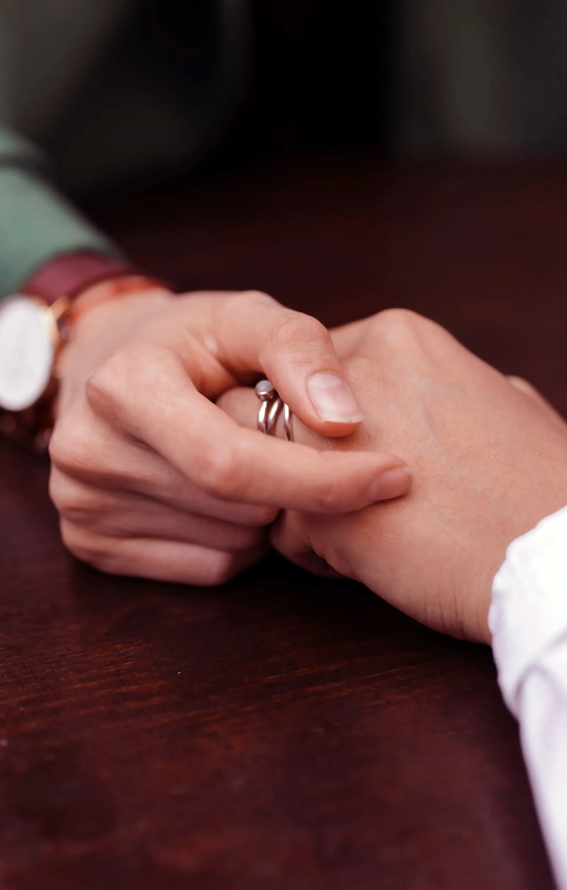 Two hands held together over a dark wooden surface; one wrist wears a watch, and fingers have thin silver rings.