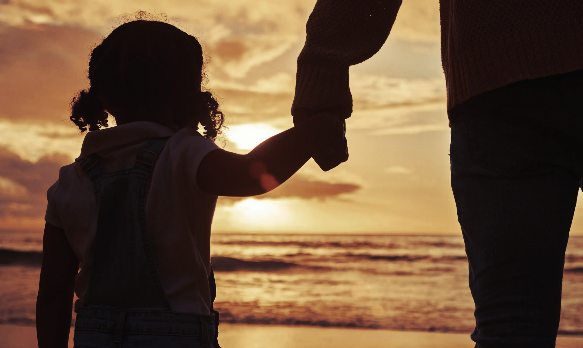 A silhouetted adult and child hold hands on a beach during a golden sunset, looking out toward the ocean.