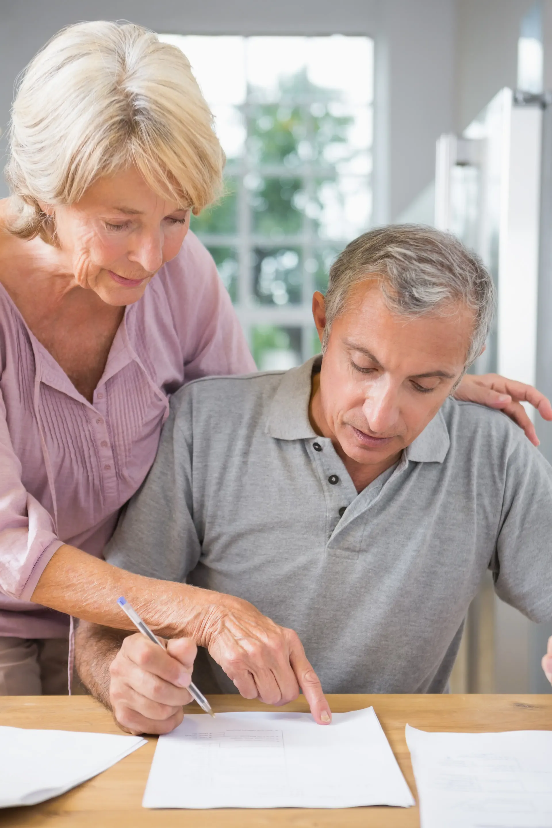 Two people look closely at documents on a wooden table, one person pointing to a paper as the other holds a pen.