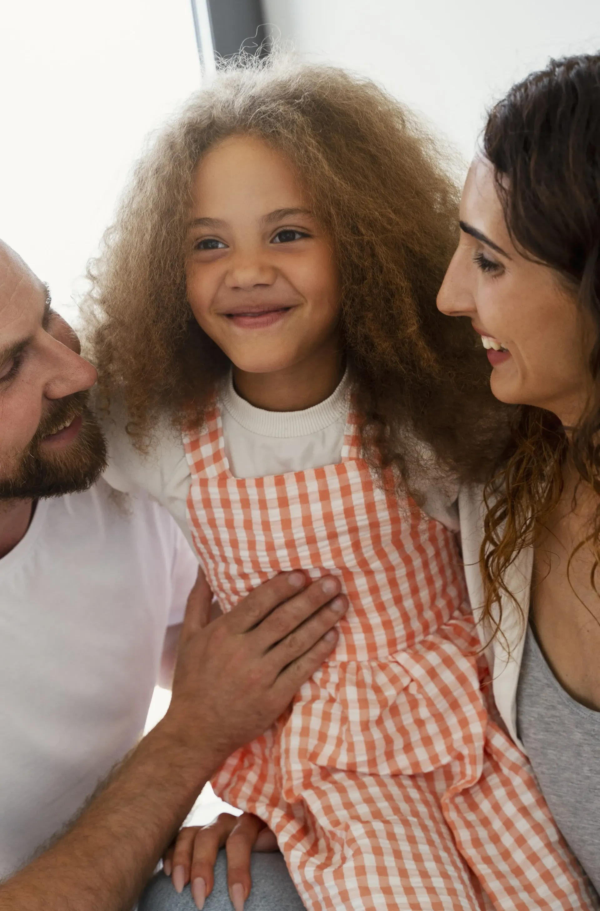 A young child with curly hair wearing orange gingham overalls sits between two adults who are smiling at them.