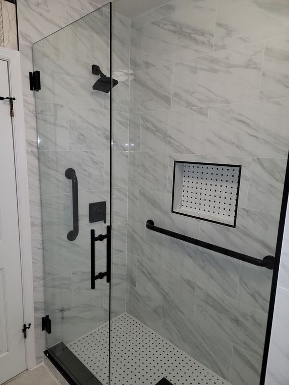 Walk-in shower with glass door, marble tile walls, black grab bars, and a fold-down shower seat