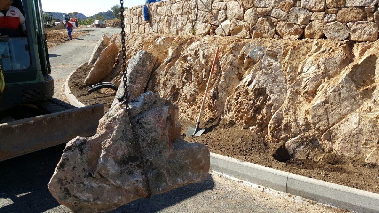 Una grande roccia viene sollevata da una gru davanti a un muro di pietra.