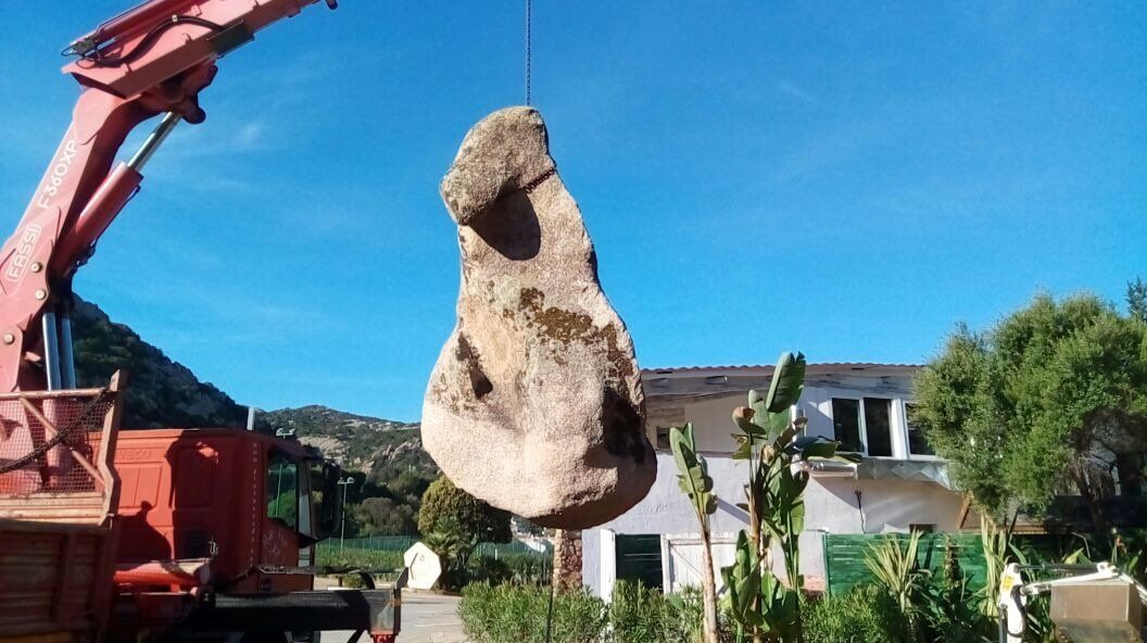 Una grande roccia viene sollevata da una gru davanti a una casa.