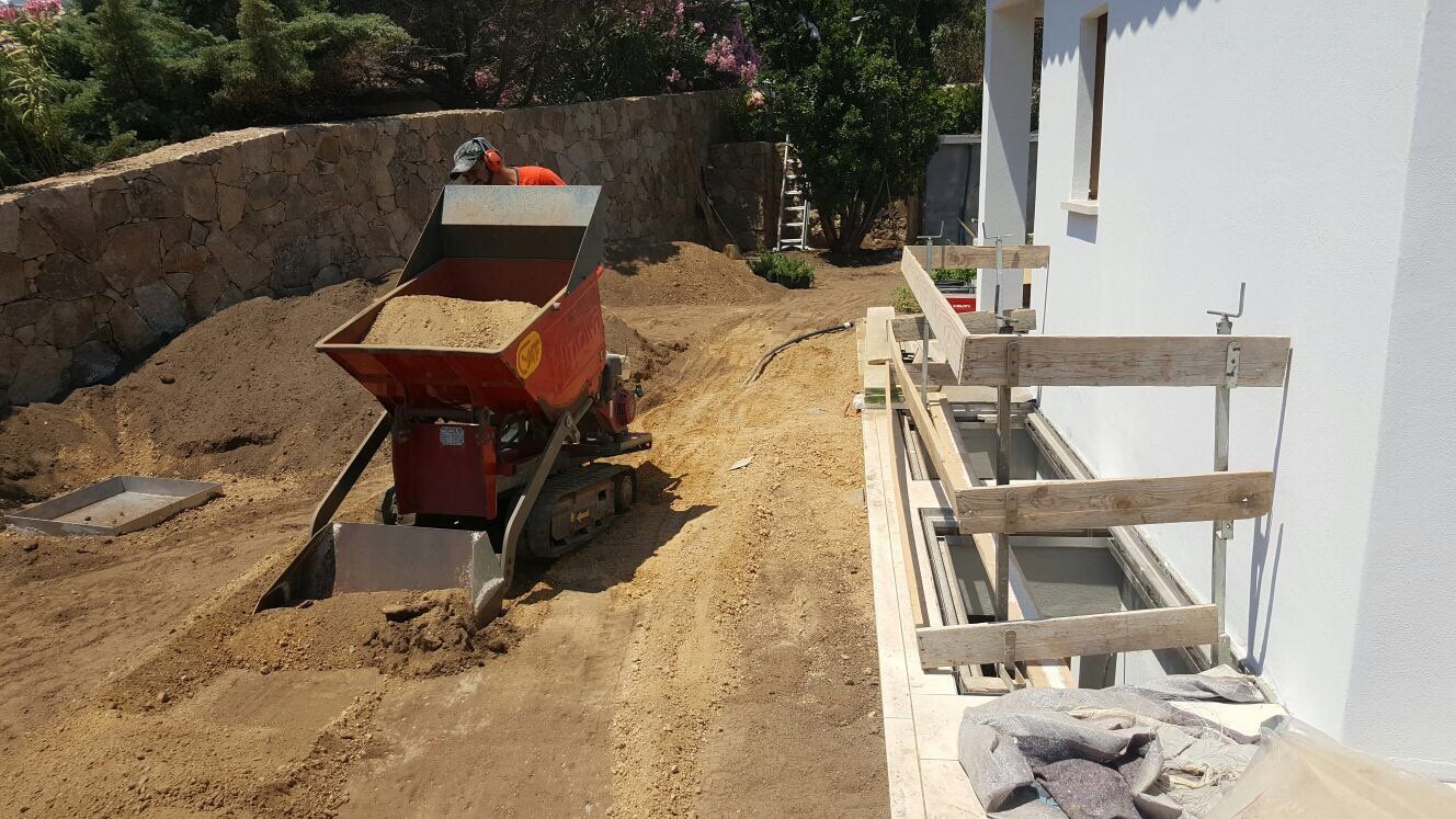 Un autocarro con cassone ribaltabile rosso è pieno di terra davanti a un edificio bianco.