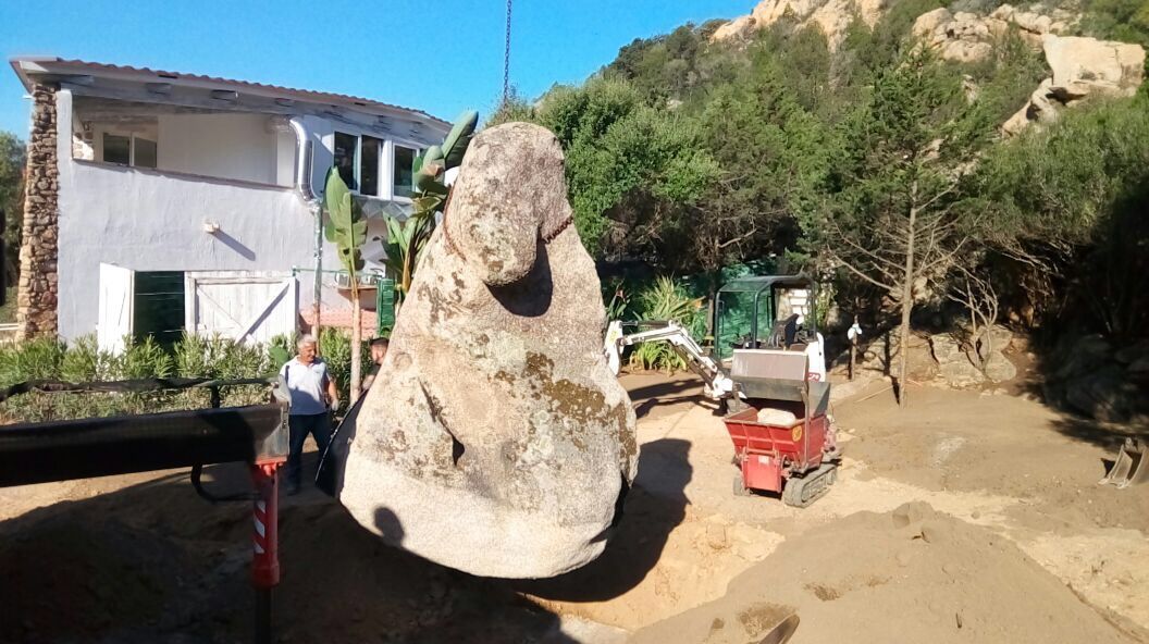 A large rock is lifted by a crane in front of a house.