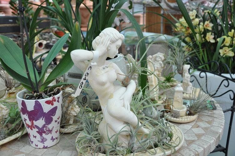 Su un tavolo circondato da piante in vaso è seduta la statua di una donna.