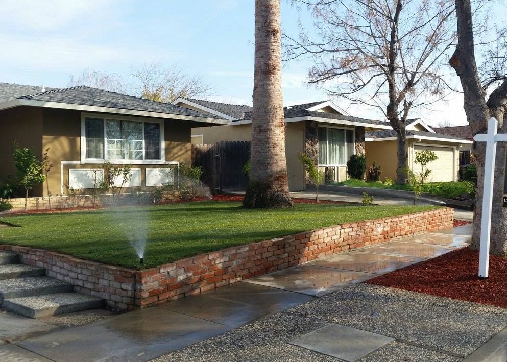 A lawn sprinkler is spraying water on a lush green lawn in front of a house.