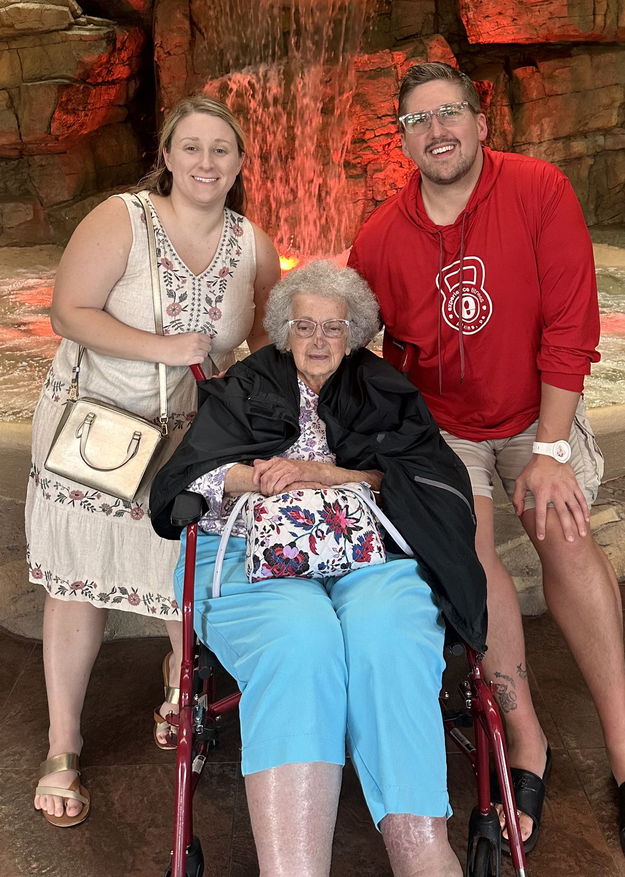Three people pose near a waterfall: woman in wheelchair, woman in dress, man in red hoodie.