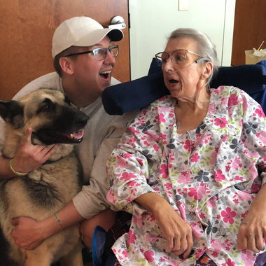 Man, senior woman, and dog in a room. Man hugs dog and looks at woman. Woman in floral top looks at man, mouth open.