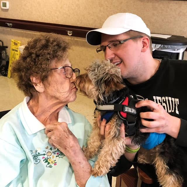 Three people pose with a sign and balloons. One person in a purple headscarf and shirt holds a sign. Two others smile.