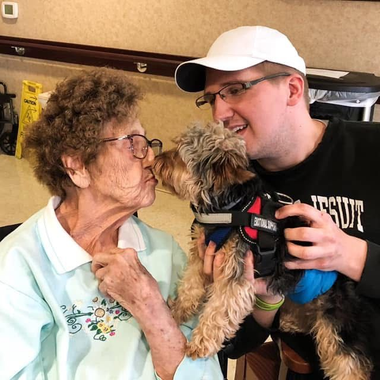 Three people pose with a sign and balloons. One person in a purple headscarf and shirt holds a sign. Two others smile.
