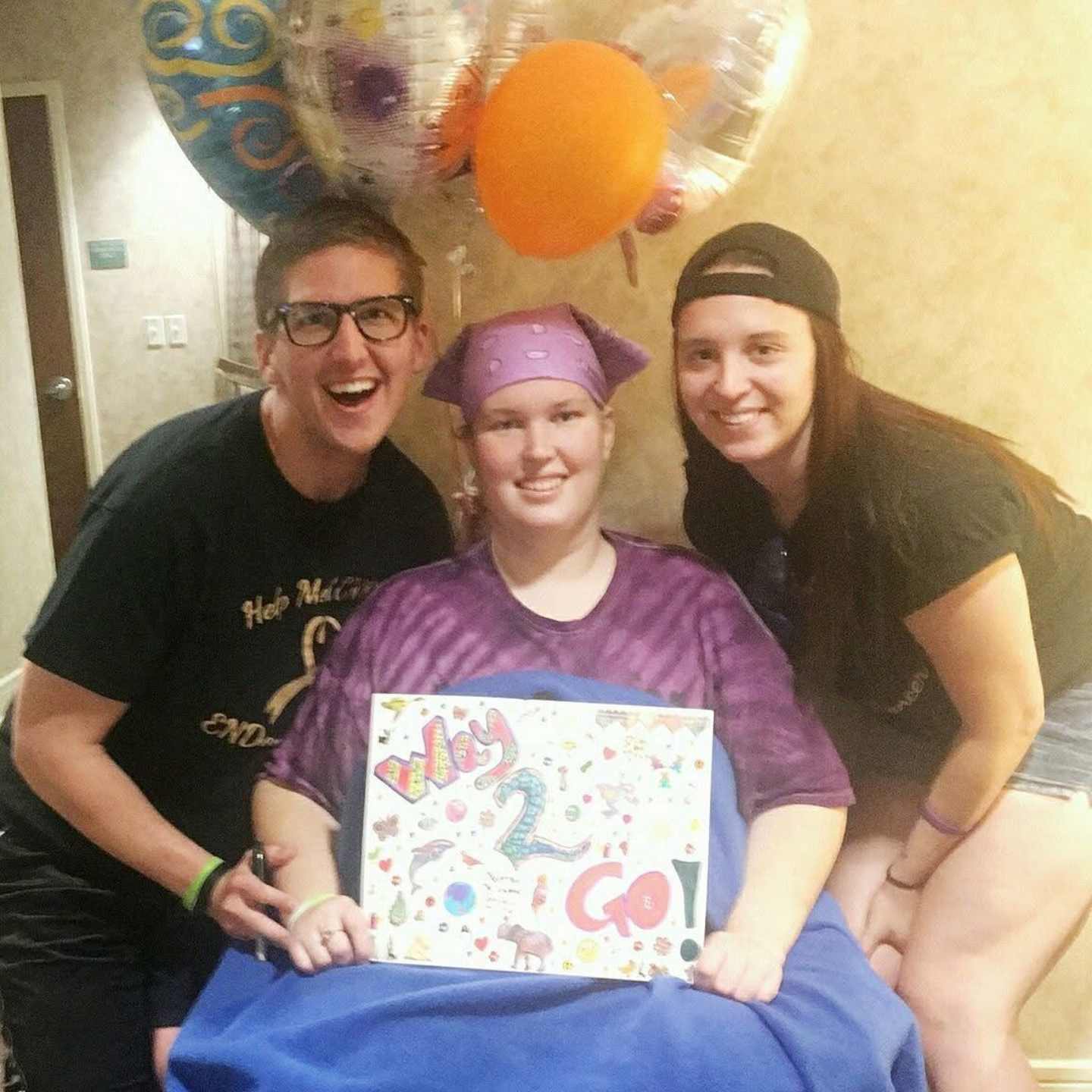 Three people pose with a sign and balloons. One person in a purple headscarf and shirt holds a sign. Two others smile.