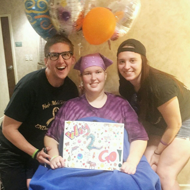 Three people pose with a sign and balloons. One person in a purple headscarf and shirt holds a sign. Two others smile.