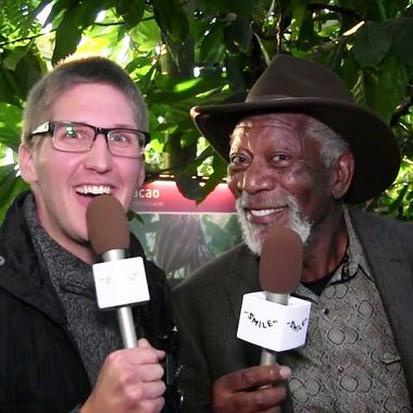 Man with glasses and Morgan Freeman holding microphones, smiling in front of greenery.